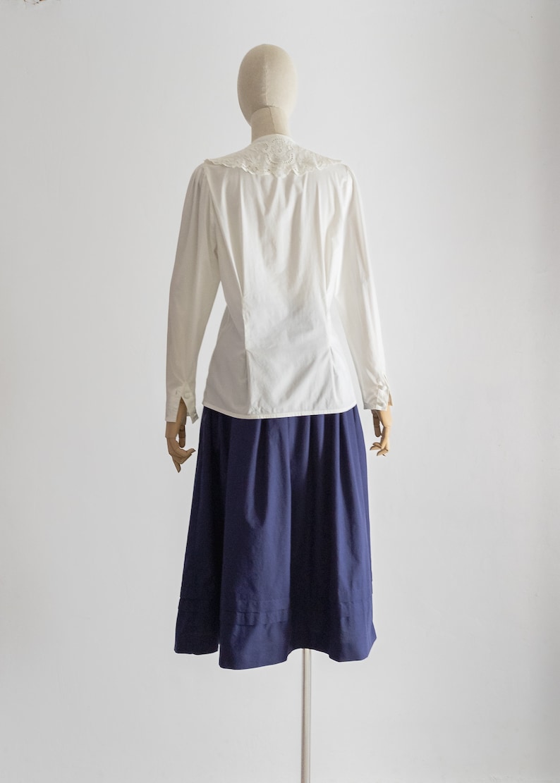 May include: A white blouse with a lace collar and long sleeves, paired with a navy blue skirt. The skirt is a knee-length A-line design. The outfit is displayed on a mannequin, showcasing a classic style.
