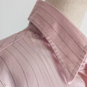 May include: Close-up of a pink button-down shirt with thin, vertical black pinstripes. The shirt is made of a smooth, possibly satin-like fabric. The collar is visible, and the shirt appears to be on a mannequin.