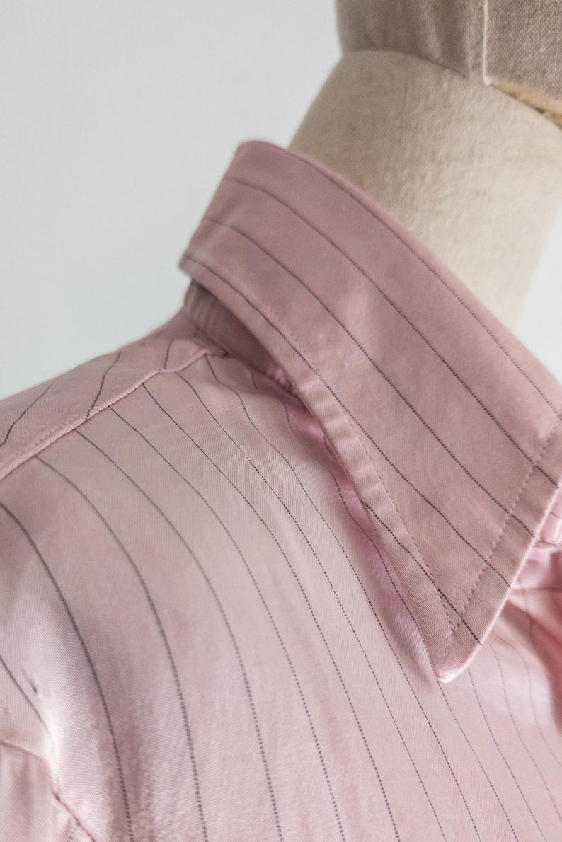 May include: Close-up of a pink button-down shirt with thin, vertical black pinstripes. The shirt is made of a smooth, possibly satin-like fabric. The collar is visible, and the shirt appears to be on a mannequin.