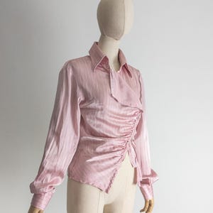 May include: A light pink long-sleeved blouse with vertical stripes and a unique asymmetrical design. The blouse features a collared neckline and ruched detailing on the front. The sleeves have buttoned cuffs. The blouse is displayed on a mannequin.