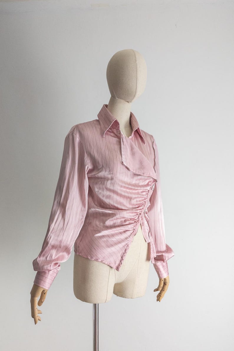 May include: A light pink long-sleeved blouse with vertical stripes and a unique asymmetrical design. The blouse features a collared neckline and ruched detailing on the front. The sleeves have buttoned cuffs. The blouse is displayed on a mannequin.
