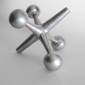 K&ouml;nnte beinhalten: Eine silberfarbene Metallskulptur in Form eines Spiel-Jacks, bestehend aus einem zentralen Knotenpunkt mit sechs strahlenf&ouml;rmigen Armen, die jeweils in einer glatten, kugelf&ouml;rmigen Kugel enden. Das Objekt hat eine matte, metallische Oberfl&auml;che.