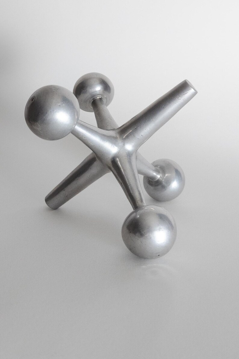 K&ouml;nnte beinhalten: Eine silberfarbene Metallskulptur in Form eines Spiel-Jacks, bestehend aus einem zentralen Knotenpunkt mit sechs strahlenf&ouml;rmigen Armen, die jeweils in einer glatten, kugelf&ouml;rmigen Kugel enden. Das Objekt hat eine matte, metallische Oberfl&auml;che.