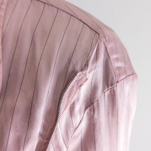 May include: Close-up of a pink jacket with vertical black pinstripes. The jacket features a silver zipper and a pink fabric detail on the shoulder. The fabric has a slight sheen.
