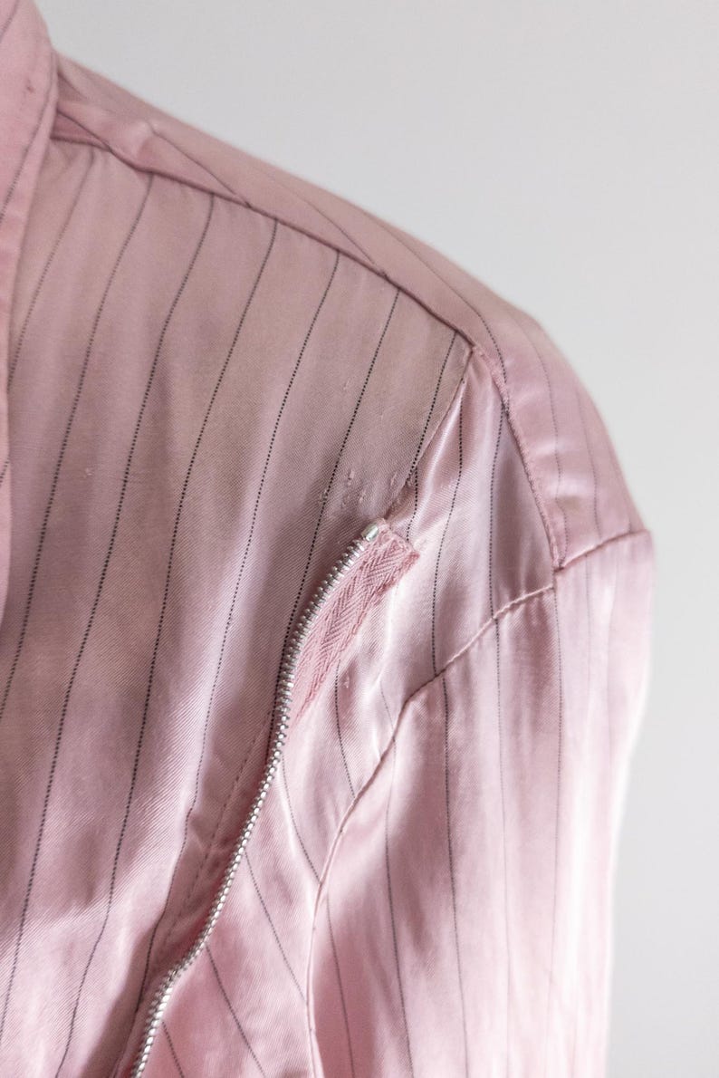 May include: Close-up of a pink jacket with vertical black pinstripes. The jacket features a silver zipper and a pink fabric detail on the shoulder. The fabric has a slight sheen.