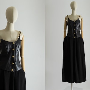 May include: A black leather corset top with gold chain straps and a row of gold buttons down the front. The top is paired with black wide-leg pants.