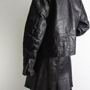 May include: A black leather jacket with a matching skirt. The jacket has a classic biker style with a notched collar, shoulder epaulets, and zippered sleeves. The skirt is a mini-skirt. The outfit is worn with black leggings.