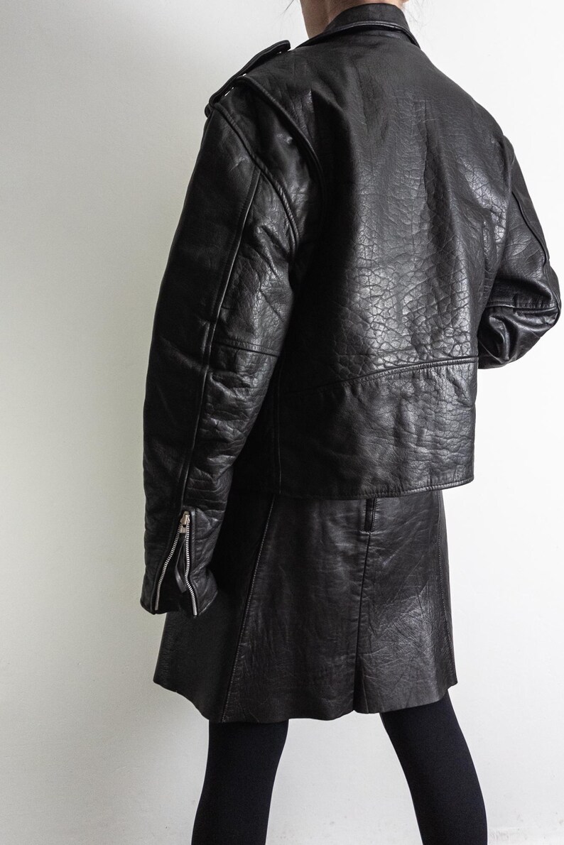 May include: A black leather jacket with a matching skirt. The jacket has a classic biker style with a notched collar, shoulder epaulets, and zippered sleeves. The skirt is a mini-skirt. The outfit is worn with black leggings.