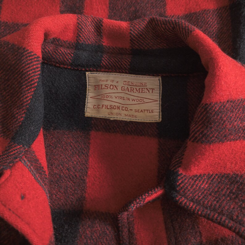May include: Close-up of a red and black checked flannel shirt with a label that reads "This is a Genuine Filson Garment 100% Virgin Wool C.C. Filson Co. - Seattle Union Made".