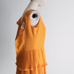 May include: An orange sleeveless dress with a pleated skirt, displayed on a mannequin. The dress has a fitted bodice and a tiered, ruffled skirt. The fabric appears to be a lightweight material, and the dress is a vibrant shade of orange.
