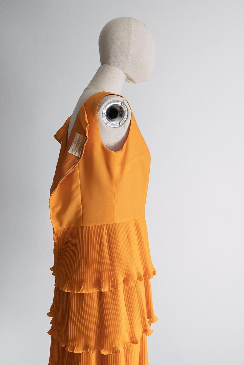 May include: An orange sleeveless dress with a pleated skirt, displayed on a mannequin. The dress has a fitted bodice and a tiered, ruffled skirt. The fabric appears to be a lightweight material, and the dress is a vibrant shade of orange.