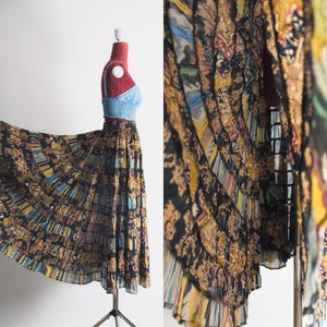 Può includere: Gonna lunga fantasia nei toni del nero, marrone, giallo e blu, esposta su un manichino. La gonna ha un design fluido e una silhouette a pieghe. Il tessuto appare leggero. Il manichino indossa un top blu.