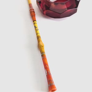 May include: A yellow and orange decorative smoking pipe with silver accents, resting on a white surface next to a red glass ashtray with a cigarette inside.