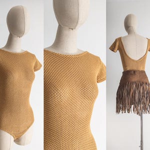 May include: A mustard-colored, short-sleeved bodysuit with a scoop neck is displayed on a mannequin. The knit fabric has a textured appearance. The bodysuit is paired with a brown suede fringe skirt.