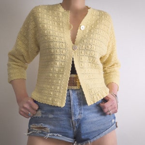 May include: A light yellow, open-front cardigan with a textured, bobble stitch design. The short-sleeved sweater has two large buttons and a scalloped edge. The cardigan is paired with denim shorts.
