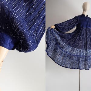 Puede incluir: Vestido azul marino con mangas largas y cuello alto. El vestido presenta una falda plisada y un corpiño con detalles de botones. La tela tiene una textura brillante con acentos plateados, creando una estética vintage. Un vestido elegante.