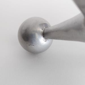K&ouml;nnte beinhalten: Ein glatter, kugelf&ouml;rmiger silberfarbener Metallknopf, der an einem konisch zulaufenden zylindrischen Metallarm befestigt ist. Die Oberfl&auml;che der Kugel zeigt einen subtilen metallischen Glanz mit kleinen Unregelm&auml;&szlig;igkeiten am Verbindungspunkt.