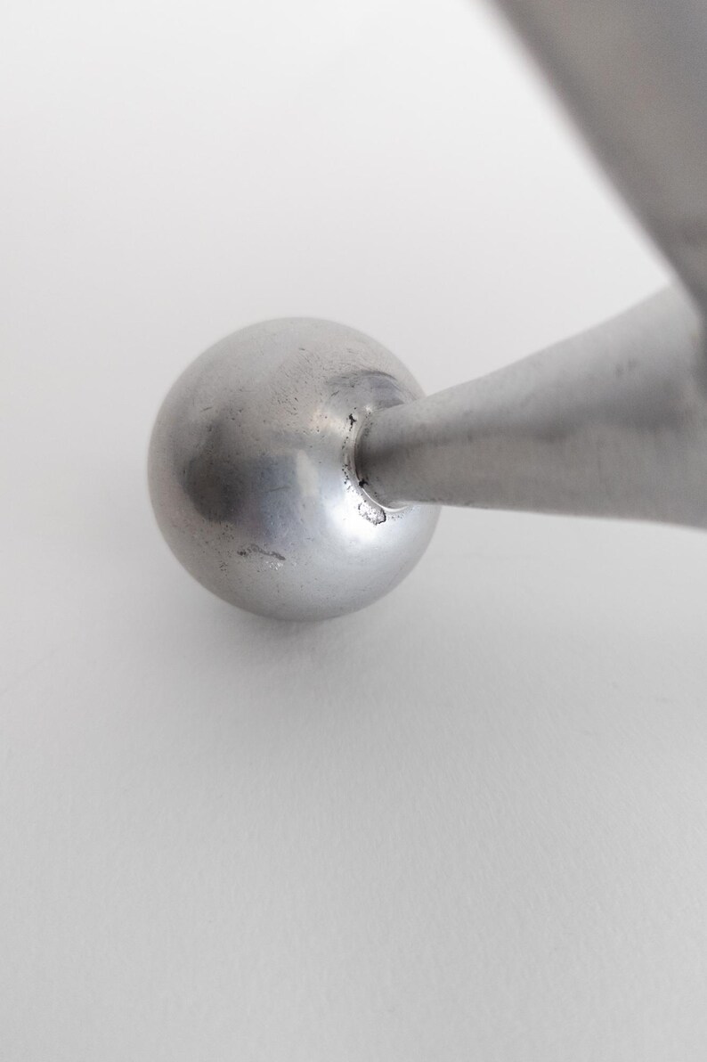 K&ouml;nnte beinhalten: Ein glatter, kugelf&ouml;rmiger silberfarbener Metallknopf, der an einem konisch zulaufenden zylindrischen Metallarm befestigt ist. Die Oberfl&auml;che der Kugel zeigt einen subtilen metallischen Glanz mit kleinen Unregelm&auml;&szlig;igkeiten am Verbindungspunkt.