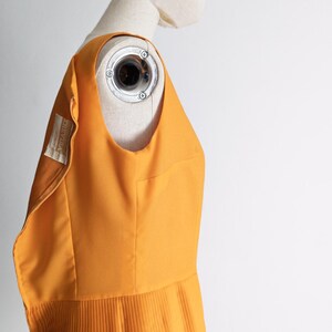 May include: A sleeveless, mustard-yellow dress with a pleated skirt is displayed on a mannequin. The dress has a scoop neckline and a fitted bodice. A label with text is visible on the dress. The background is plain white.
