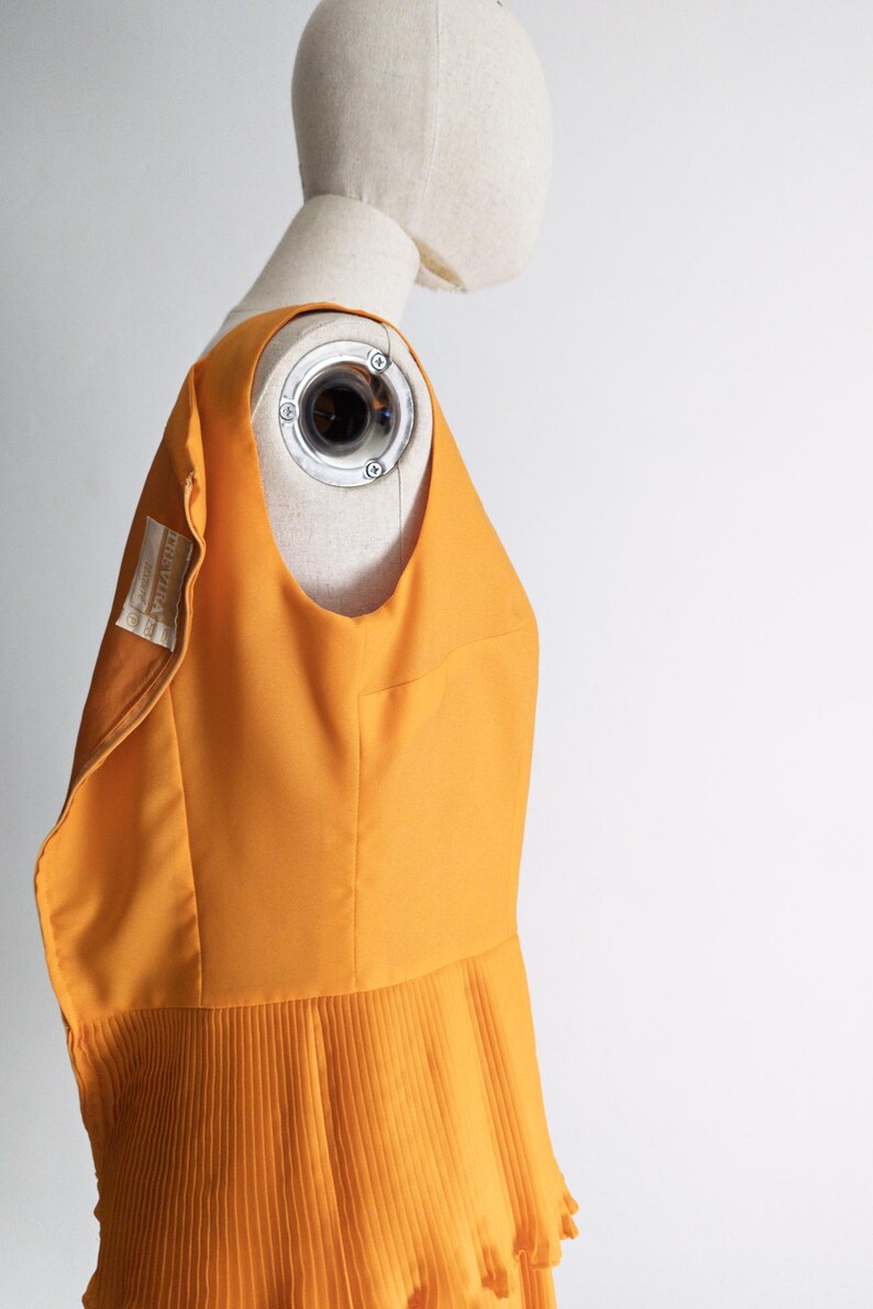 May include: A sleeveless, mustard-yellow dress with a pleated skirt is displayed on a mannequin. The dress has a scoop neckline and a fitted bodice. A label with text is visible on the dress. The background is plain white.