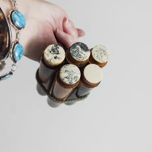 Puede incluir: Colección de seis pequeñas botellas de vidrio vintage con tapas decorativas, sujetas con un cordón verde. Las tapas tienen un diseño veteado en tonos negro, blanco y crema. Se ve un brazalete de plata con piedras turquesas.