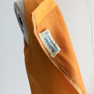 May include: Close-up of a sleeveless, orange dress on a mannequin. The dress has a zipper detail and a label that reads "MADRISA Irene Modell". The fabric appears smooth and the dress is a solid color.