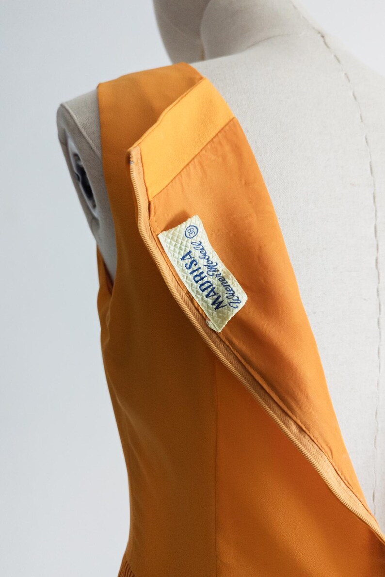 May include: Close-up of a sleeveless, orange dress on a mannequin. The dress has a zipper detail and a label that reads "MADRISA Irene Modell". The fabric appears smooth and the dress is a solid color.