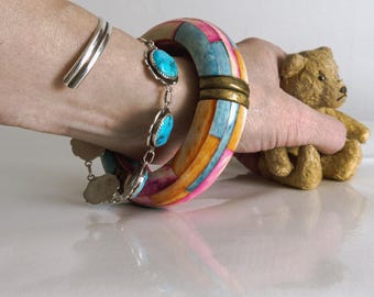 May include: A vibrant, chunky bracelet with turquoise detailing and a silver cuff. The large bracelet has a mosaic design with pink, blue, and orange segments. A small teddy bear is held in the background.