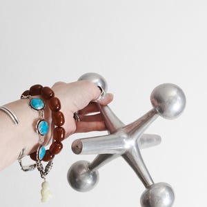 K&ouml;nnte beinhalten: Eine gro&szlig;e, silberfarbene Metallskulptur in Form eines Spiel-Jacks mit sechs abgerundeten Spitzen. Das Objekt hat eine glatte, reflektierende Oberfl&auml;che und eine sternf&ouml;rmige geometrische Struktur mit gleichm&auml;&szlig;igen Armen, die von einem Kern ausgehen.