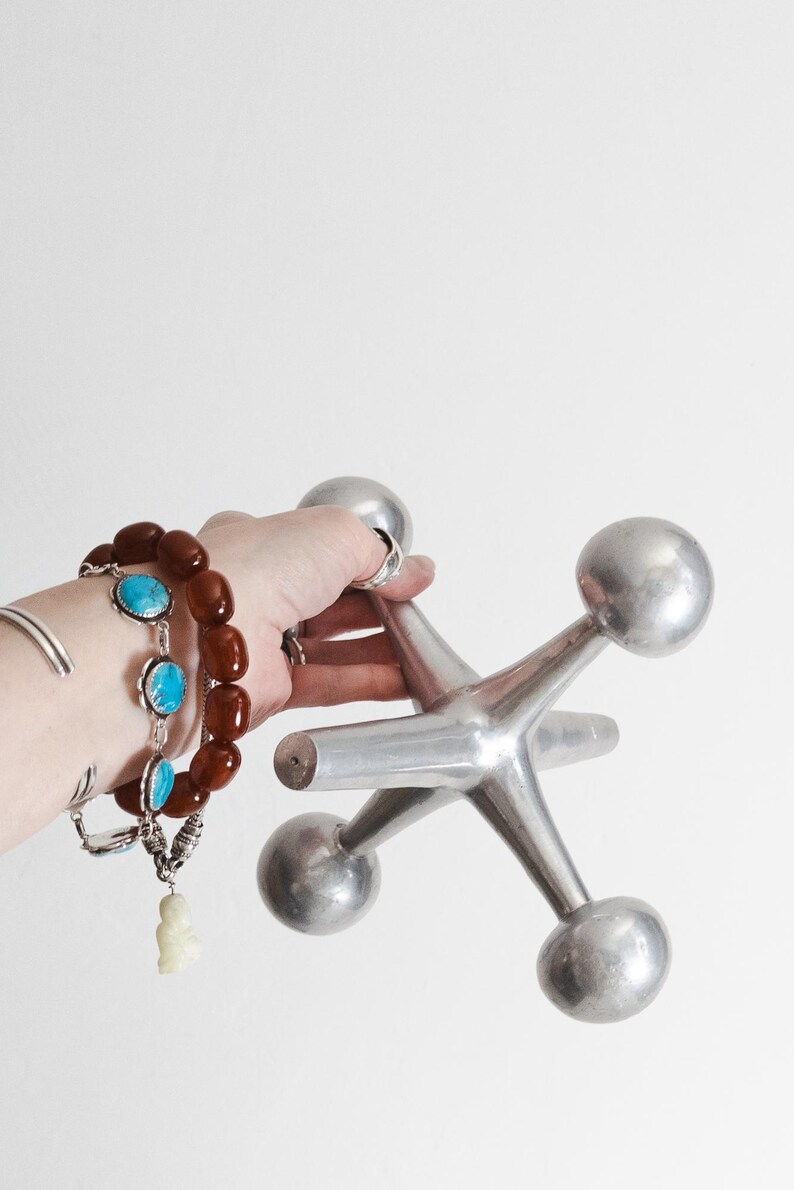 K&ouml;nnte beinhalten: Eine gro&szlig;e, silberfarbene Metallskulptur in Form eines Spiel-Jacks mit sechs abgerundeten Spitzen. Das Objekt hat eine glatte, reflektierende Oberfl&auml;che und eine sternf&ouml;rmige geometrische Struktur mit gleichm&auml;&szlig;igen Armen, die von einem Kern ausgehen.