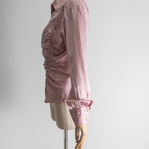 May include: A light pink button-down shirt with vertical stripes and a ruched front. The long-sleeved shirt has a classic collar, buttoned cuffs, and a zipper detail on the shoulder. The shirt is displayed on a mannequin.
