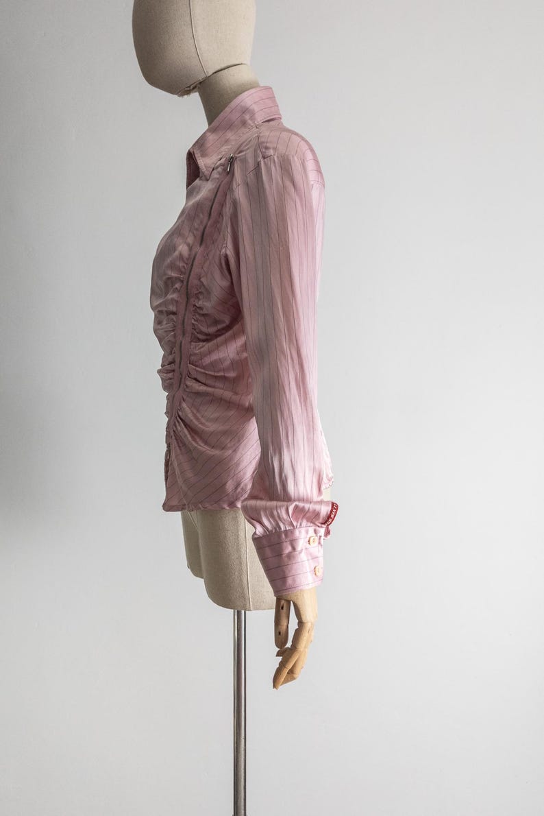 May include: A light pink button-down shirt with vertical stripes and a ruched front. The long-sleeved shirt has a classic collar, buttoned cuffs, and a zipper detail on the shoulder. The shirt is displayed on a mannequin.