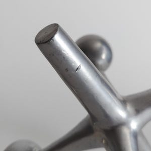K&ouml;nnte beinhalten: Eine silberfarbene Metallskulptur in Form eines Spiel-Jacks mit sternf&ouml;rmigen, in abgerundeten Kugeln endenden Armen. Die Metalloberfl&auml;che weist ein glattes, mattes Finish auf, mit einem kleinen, sichtbaren Kratzer an einem der zentralen Arme.