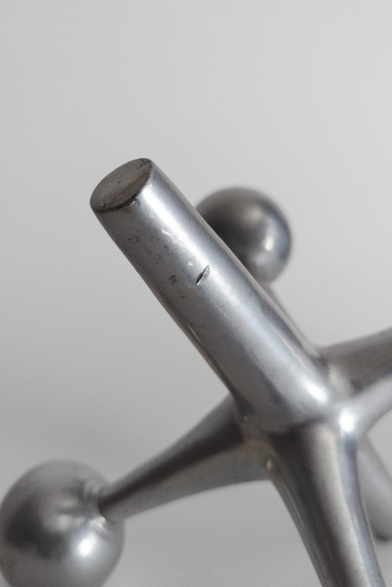 K&ouml;nnte beinhalten: Eine silberfarbene Metallskulptur in Form eines Spiel-Jacks mit sternf&ouml;rmigen, in abgerundeten Kugeln endenden Armen. Die Metalloberfl&auml;che weist ein glattes, mattes Finish auf, mit einem kleinen, sichtbaren Kratzer an einem der zentralen Arme.