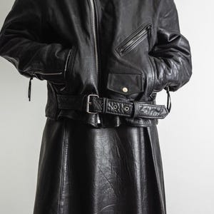 May include: Black leather jacket with a belted waist and a matching black leather skirt. The jacket features a zipper closure, multiple pockets, and shoulder epaulets. The skirt has a flared design.