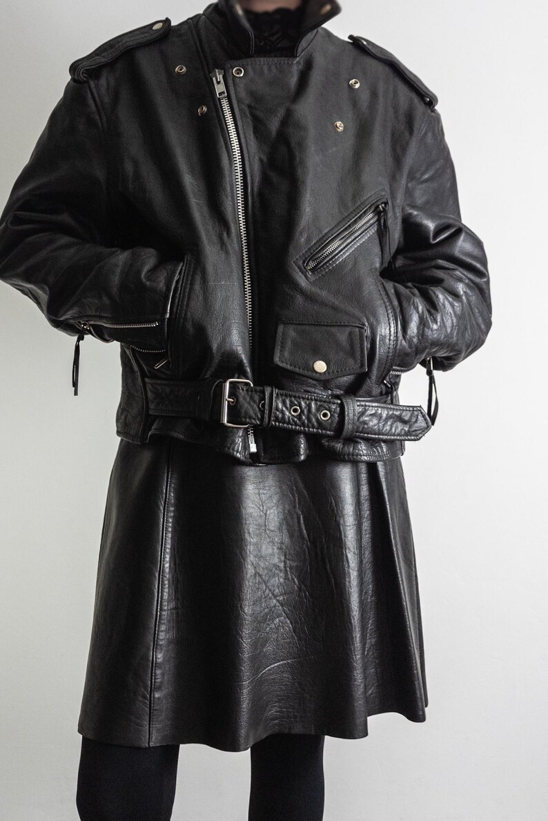 May include: Black leather jacket with a belted waist and a matching black leather skirt. The jacket features a zipper closure, multiple pockets, and shoulder epaulets. The skirt has a flared design.