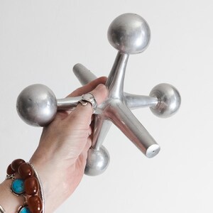 K&ouml;nnte beinhalten: Ein gro&szlig;es, silberfarbenes Metall-Spielzeug in Form eines Springers mit sechs strahlenf&ouml;rmigen Armen, die in glatten, abgerundeten Kugeln enden. Das Objekt hat eine matte, metallische Oberfl&auml;che und eine geometrische Struktur.