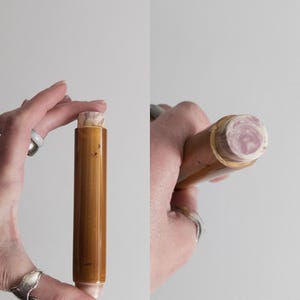 Puede incluir: Un tapón de botella cilíndrico marrón con una parte superior clara y estampada. El tapón se sostiene en una mano sobre un fondo liso. La parte superior del tapón tiene un remolino rosa y blanco.
