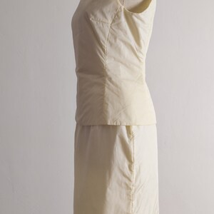 May include: A sleeveless, ivory-colored dress with a fitted bodice and a skirt that falls to the knee. The dress is made of a smooth, silky fabric.