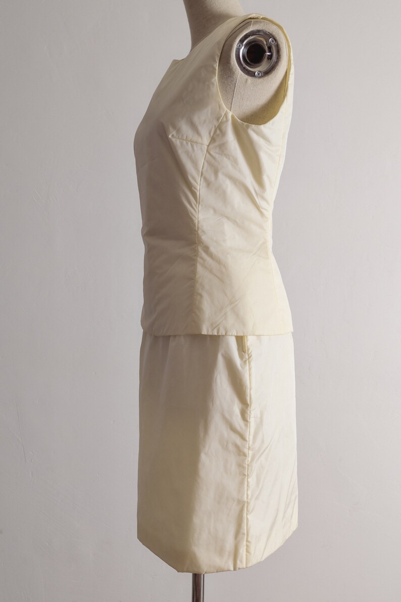 May include: A sleeveless, ivory-colored dress with a fitted bodice and a skirt that falls to the knee. The dress is made of a smooth, silky fabric.