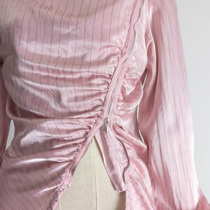 May include: A light pink, long-sleeved blouse with vertical black pinstripes. The blouse features a collared neckline and a diagonal silver zipper closure. The fabric has a silky sheen and is gathered at the waist.