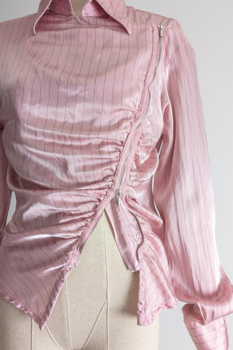 May include: A light pink, long-sleeved blouse with vertical black pinstripes. The blouse features a collared neckline and a diagonal silver zipper closure. The fabric has a silky sheen and is gathered at the waist.