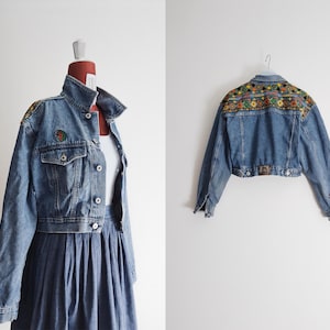 May include: A light-wash denim jacket with a colourful embroidered design on the shoulders and back. The jacket features a classic collar, button-up front, and pockets. A colourful patch is visible on the left shoulder. The jacket is paired with a denim skirt.