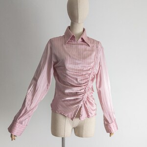May include: A light pink long-sleeve blouse with vertical stripes and a ruched side detail. The blouse has a collared neckline and buttoned cuffs. The garment is displayed on a mannequin.