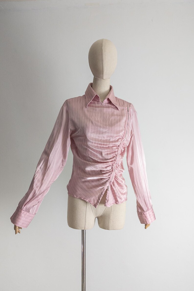 May include: A light pink long-sleeve blouse with vertical stripes and a ruched side detail. The blouse has a collared neckline and buttoned cuffs. The garment is displayed on a mannequin.