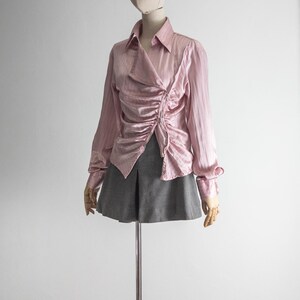 May include: A light pink, long-sleeved blouse with a ruched front detail and a collared neckline. The blouse is paired with a gray skirt. The clothing is displayed on a mannequin.