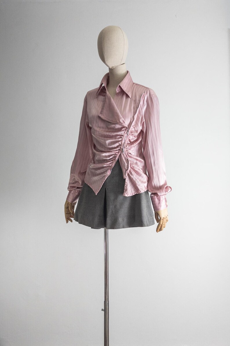 May include: A light pink, long-sleeved blouse with a ruched front detail and a collared neckline. The blouse is paired with a gray skirt. The clothing is displayed on a mannequin.