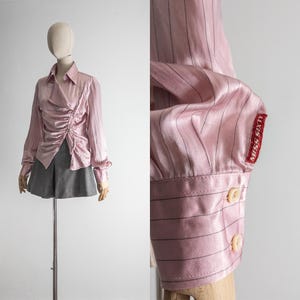 May include: A light pink satin blouse with vertical black stripes and a ruched front. The long-sleeved shirt has a collared neckline and is paired with gray shorts. The sleeve cuff features the text "MISS SIXTY".