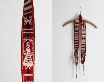 Op de afbeelding: Een lange, geweven textielriem met een herhalend patroon in rood, wit en zwart. Het ontwerp bevat gestileerde figuren van mensen en dieren, geometrische vormen en horizontale strepen. De uiteinden van de riem zijn afgewerkt met gevlochten kwastjes.