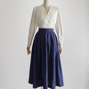May include: A vintage outfit displayed on a mannequin. It comprises a white blouse with a lace collar and long sleeves, and a navy blue pleated midi skirt. The outfit is set against a plain white backdrop.